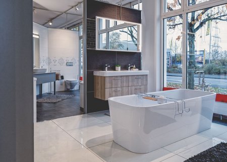 Großzügige Badausstellung mit freistehender Badewanne, Doppelwaschtisch, großem Spiegel und bodentiefen Fenstern mit Blick nach draußen.