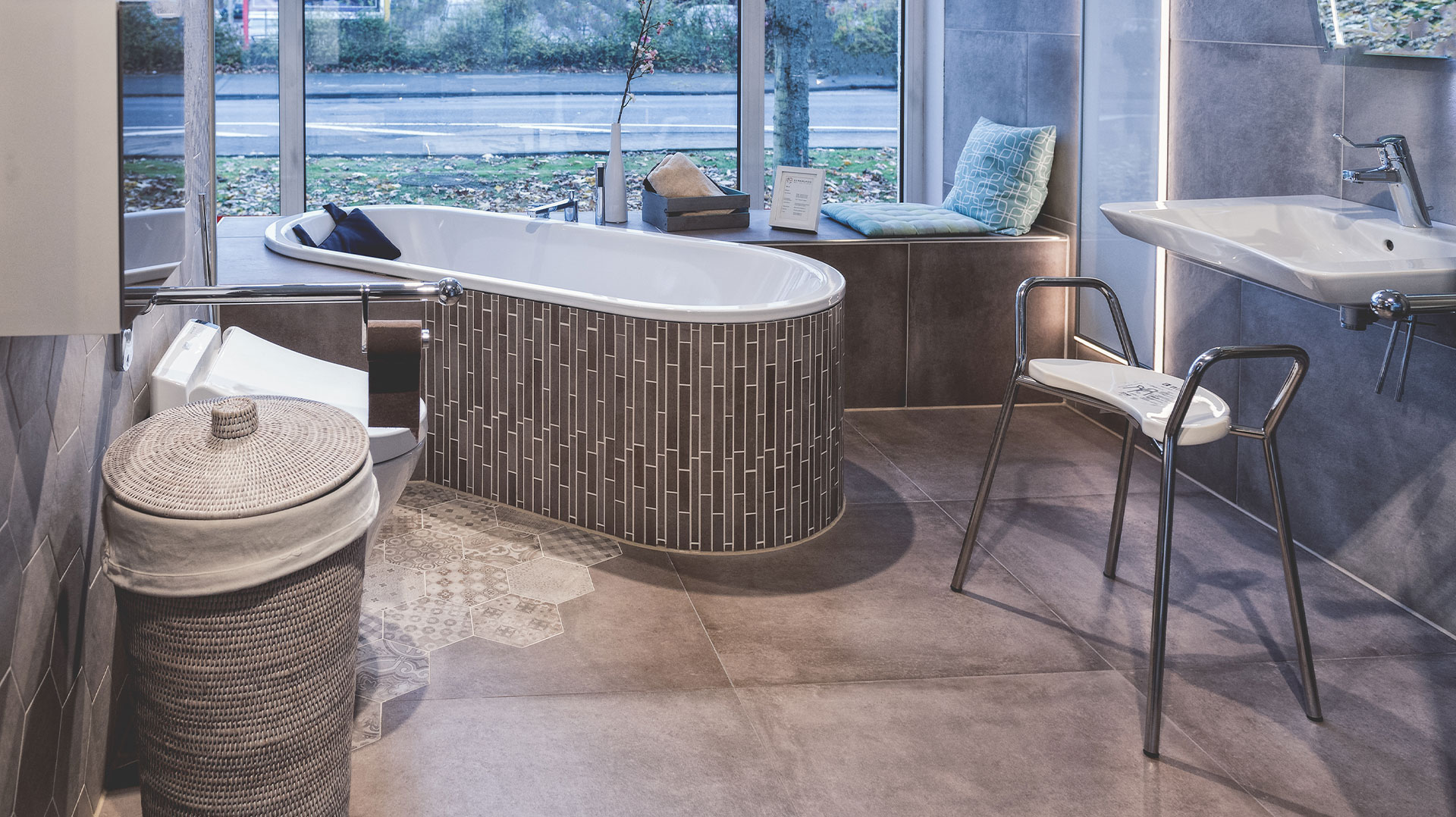 Ausstellung eines barrierefreien Badezimmers mit niedrig zugänglicher Badewanne, Haltegriffen, Waschbecken mit Stuhl, grauen Fliesen und großer Fensterfront.